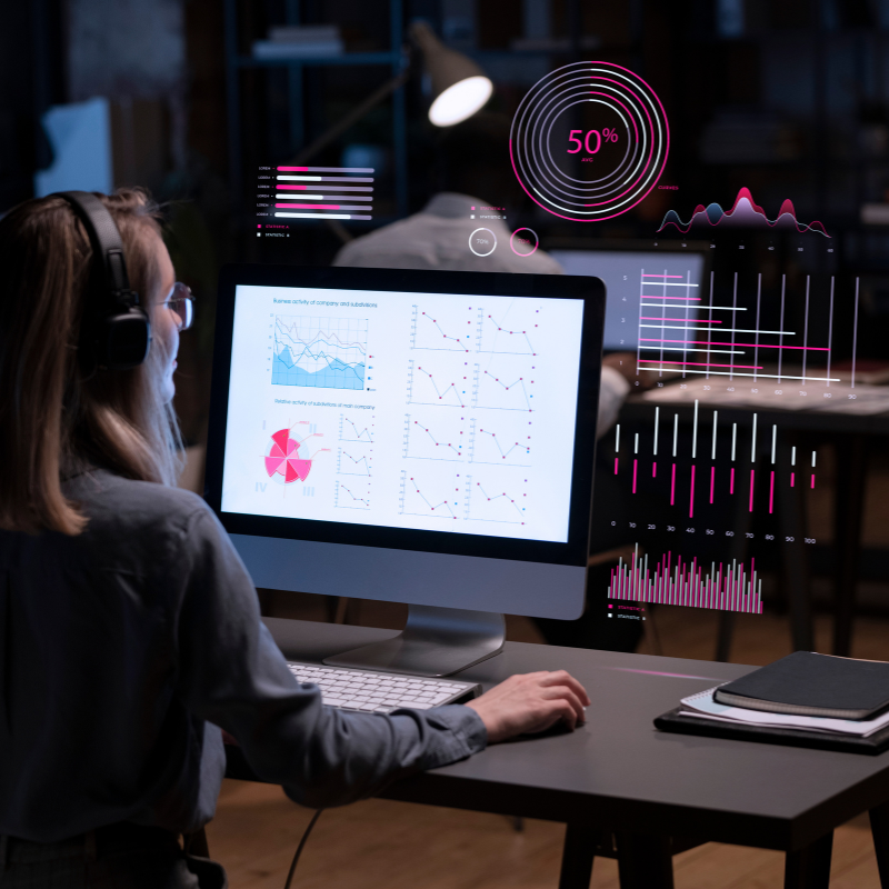 Mulher de fones de ouvido sentada em uma mesa no escritório, olhando para o monitor do computador, onde gráficos e dados de marketing digital são exibidos em realidade aumentada ao redor dela.