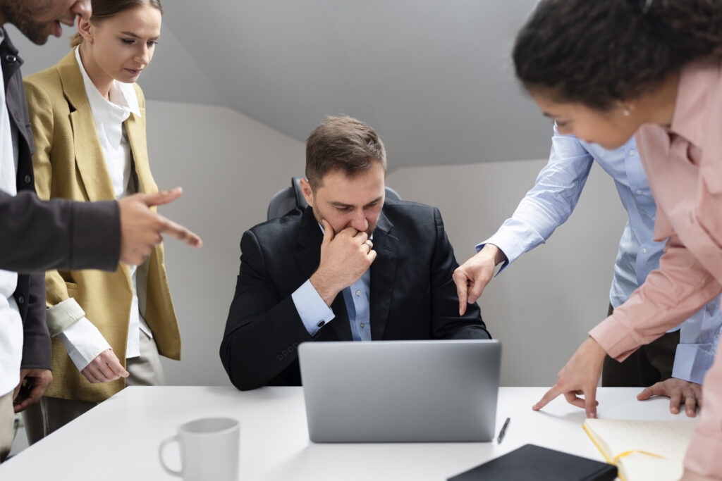 
Um homem de meia-idade, sentado em uma mesa de escritório, olha para baixo com uma expressão de preocupação e insegurança. Ele está cercado por quatro colegas, cujos rostos não são totalmente visíveis, mas que estão apontando dedos para ele e para o laptop em sua mesa. A cena sugere um momento de assédio ou crítica severa no ambiente de trabalho.