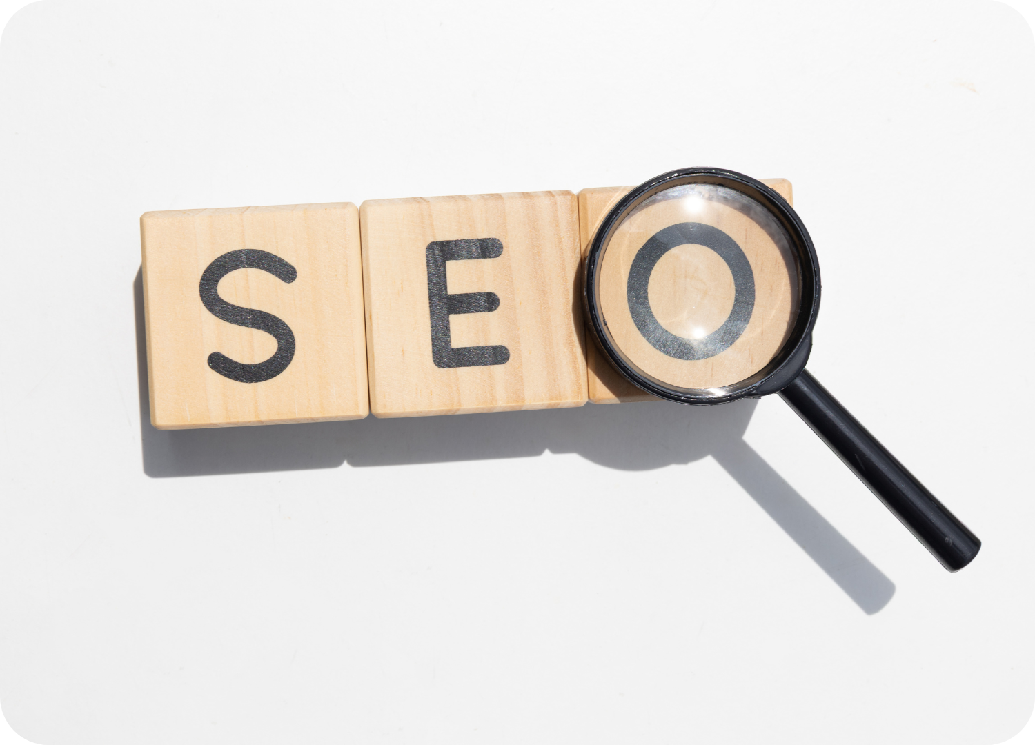 Blocos de madeira formando a sigla SEO, com uma lupa amplificando a letra O. A imagem simboliza a otimização de sites e a busca por resultados.