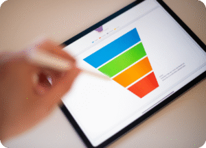 Mão de uma pessoa segurando uma caneta digital (stylus) e apontando para um gráfico de funil de vendas multicolorido (azul, verde, laranja e vermelho) exibido na tela de um tablet. O gráfico representa visualmente as etapas do funil de vendas.