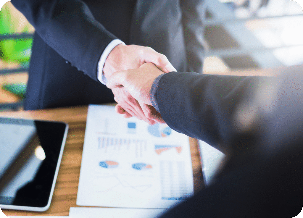 Aperto de mãos de dois executivos sobre uma mesa com relatórios e gráficos. A imagem simboliza parceria estratégica, fechamento de negócio e consultoria de marketing de resultados.