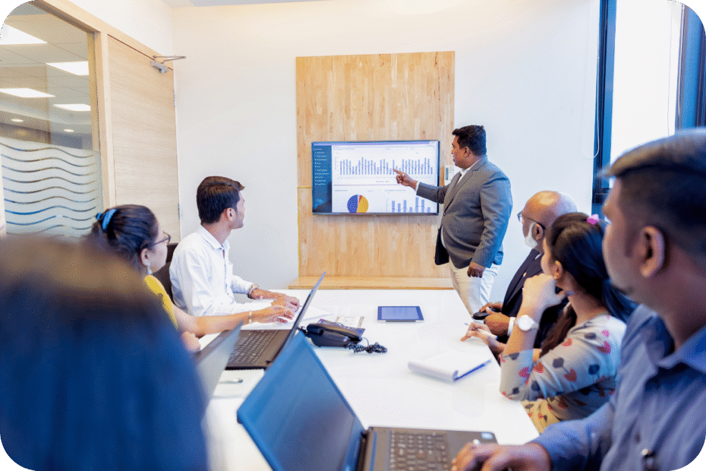 Um executivo de terno cinza, possivelmente o CMO ou líder de marketing, apresentando dados de desempenho em uma tela grande para um grupo de colegas (incluindo o CFO) em uma sala de reuniões. Ele aponta para um gráfico que demonstra KPIs financeiros, ilustrando a discussão sobre estratégia e orçamento com base em dados.