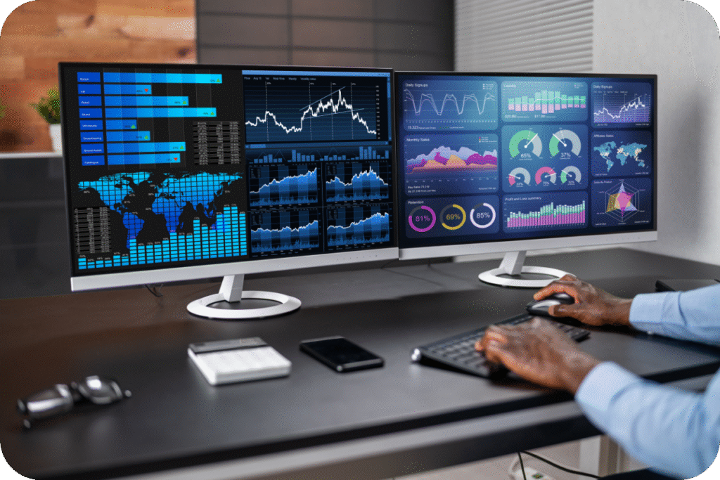 Uma pessoa sentada em uma mesa, digitando em um teclado e usando um mouse, em frente a dois monitores grandes. Ambos os monitores exibem painéis de dados (dashboards) complexos e detalhados em tons de azul, verde e branco, incluindo gráficos de barras, gráficos de linha, gráficos circulares e mapas-múndi. Os dados visíveis incluem métricas de vendas mensais, retenção e desempenho de mercado global, sugerindo análise financeira e estratégica de alto nível.