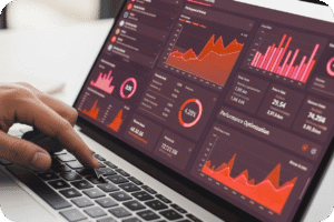 Uma foto em close-up de uma mão tocando um teclado de laptop, que exibe um painel de dados (dashboard) em tons de vermelho e cinza. O painel está cheio de gráficos e métricas de negócios, incluindo gráficos de linha, barras e um gráfico de pizza, focados em otimização de performance e KPIs financeiros, sugerindo análise de dados e marketing.