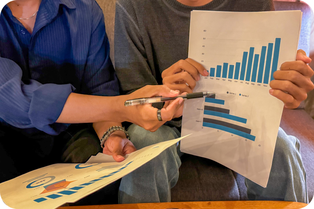 Duas pessoas analisando relatórios de desempenho impressos com gráficos de barras azuis crescentes. Uma delas aponta uma métrica específica com uma caneta, simbolizando o alinhamento estratégico e a análise conjunta de dados entre Marketing e Vendas.