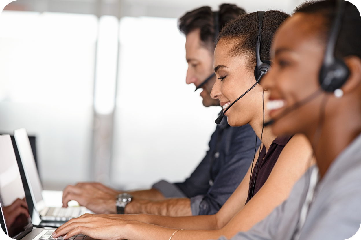 Três profissionais, um homem e duas mulheres, sentados em fila em um escritório moderno, usando headsets e sorrindo enquanto trabalham em seus laptops. A imagem representa uma equipe de Vendas e Atendimento operando de forma integrada e alinhada.