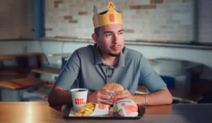 Homem jovem sentado em uma lanchonete Burger King, usando a icônica coroa de papel da marca. Ele segura um hambúrguer Whopper à frente de uma bandeja com batatas fritas e refrigerante free refill, representando a experiência de consumo em fast food.