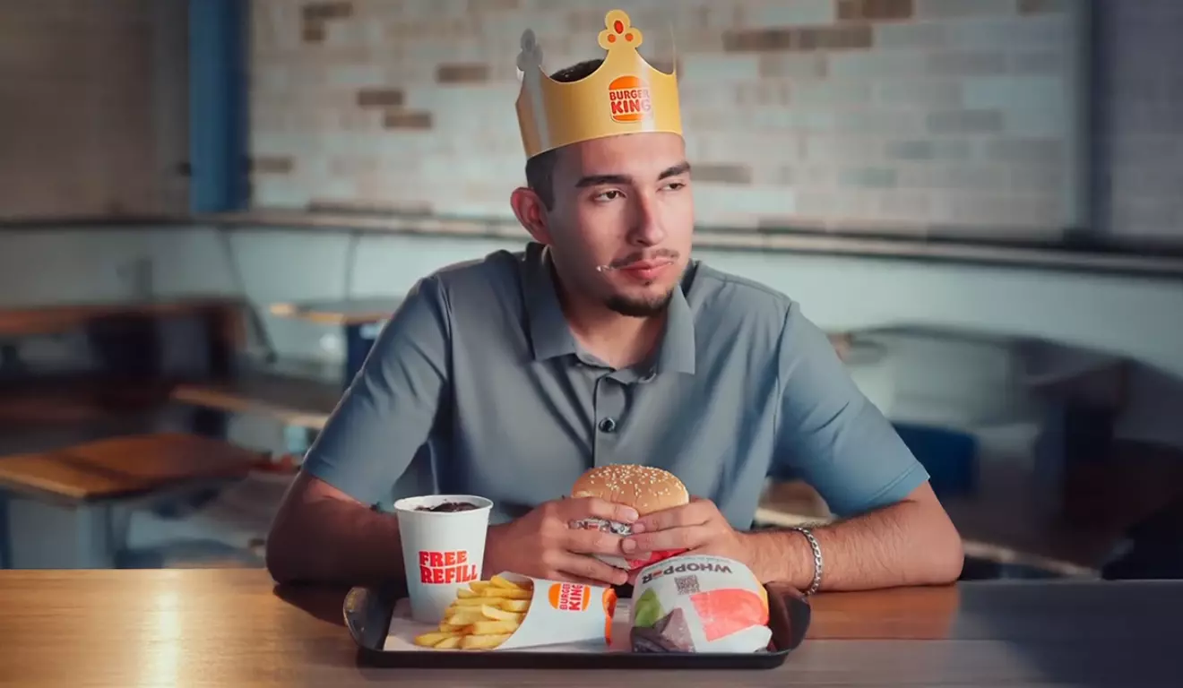 Homem jovem sentado em uma lanchonete Burger King, usando a icônica coroa de papel da marca. Ele segura um hambúrguer Whopper à frente de uma bandeja com batatas fritas e refrigerante free refill, representando a experiência de consumo em fast food.