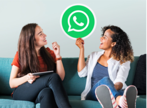 Duas jovens mulheres sorridentes sentadas em um sofá verde, uma segurando um ícone físico do WhatsApp e a outra segurando um tablet, ilustrando o uso do aplicativo para vendas e comunicação.