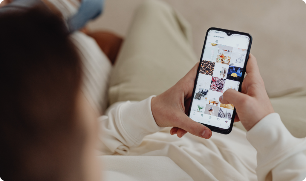 Visão sobre o ombro de uma pessoa navegando pelo feed do Instagram em um smartphone. A tela exibe uma grade de fotos estéticas, incluindo café, interiores e gráficos. A imagem ilustra o comportamento do usuário nas redes sociais, o design de interface (UI) e a curadoria de conteúdo digital para estratégias de social media marketing.