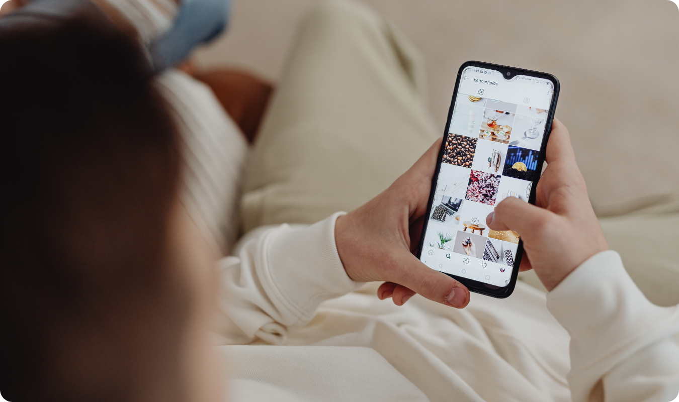 Visão sobre o ombro de uma pessoa navegando pelo feed do Instagram em um smartphone. A tela exibe uma grade de fotos estéticas, incluindo café, interiores e gráficos. A imagem ilustra o comportamento do usuário nas redes sociais, o design de interface (UI) e a curadoria de conteúdo digital para estratégias de social media marketing.