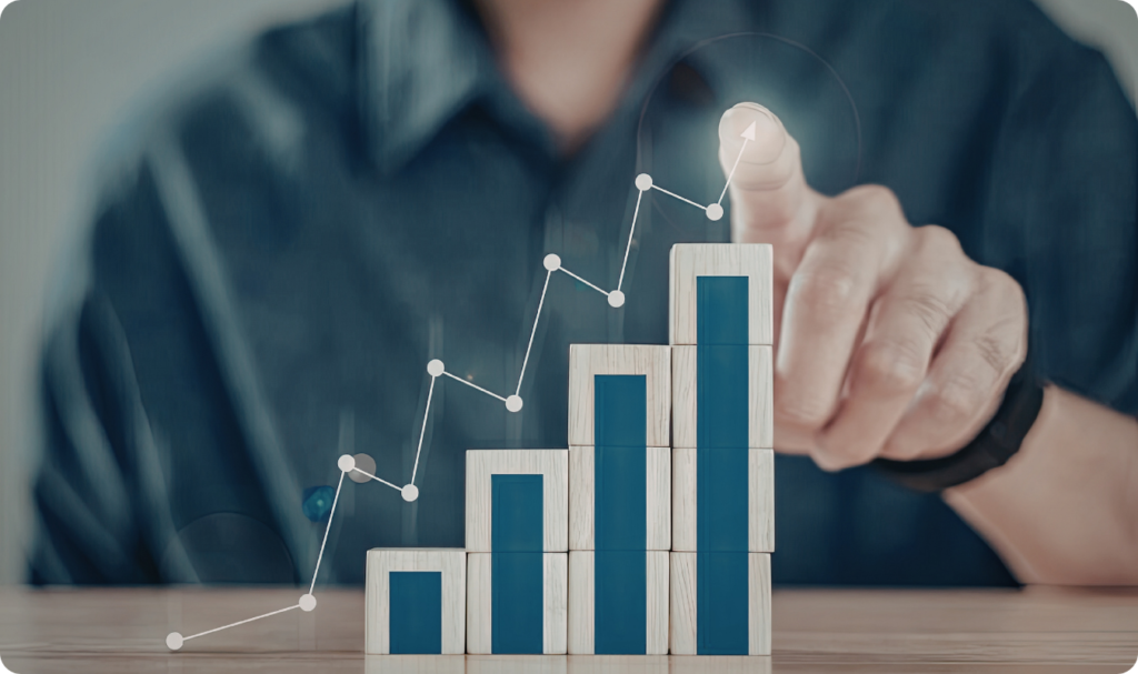 Close-up de uma mão apontando para o topo de um gráfico de barras ascendente feito de blocos de madeira. Uma linha de tendência branca com pontos de dados está sobreposta digitalmente aos blocos, simbolizando o crescimento contínuo, a análise de métricas de sucesso e a implementação de estratégias de Business Intelligence para expansão de mercado