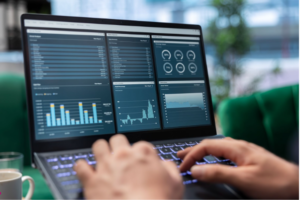 Visão aproximada de um notebook moderno exibindo um dashboard complexo de Business Intelligence. A tela mostra múltiplos gráficos de barras, indicadores de performance (KPIs) e tabelas de análise financeira em tons de azul. No primeiro plano, mãos de um profissional operam o teclado retroiluminado, simbolizando tomada de decisão baseada em dados em um ambiente corporativo.