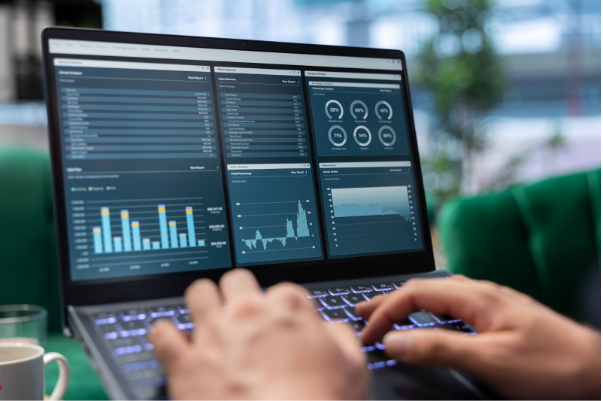 Visão aproximada de um notebook moderno exibindo um dashboard complexo de Business Intelligence. A tela mostra múltiplos gráficos de barras, indicadores de performance (KPIs) e tabelas de análise financeira em tons de azul. No primeiro plano, mãos de um profissional operam o teclado retroiluminado, simbolizando tomada de decisão baseada em dados em um ambiente corporativo.