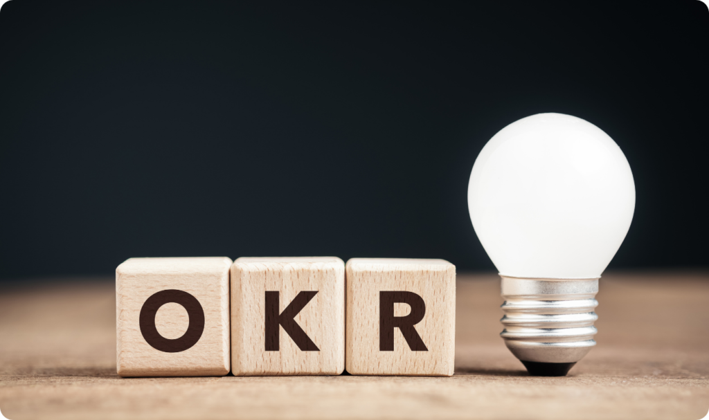 Três blocos de madeira com as letras O, K e R alinhados horizontalmente ao lado de uma lâmpada acesa sobre uma superfície de madeira, com fundo escuro. A imagem simboliza a clareza e a iluminação trazidas pela metodologia de Objetivos e Resultados-Chave.