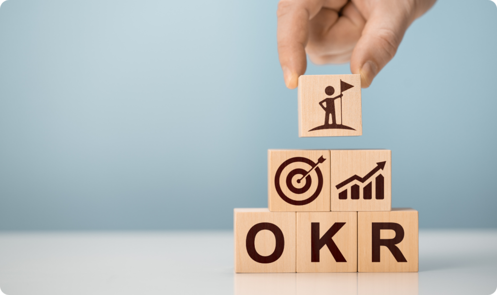 Uma mão humana coloca o último bloco de madeira no topo de uma pirâmide. Esse bloco superior exibe o ícone de uma pessoa segurando uma bandeira em um cume. Abaixo, dois blocos mostram ícones de um alvo e um gráfico de crescimento ascendente. Na base, três blocos formam a sigla OKR contra um fundo azul claro e limpo.