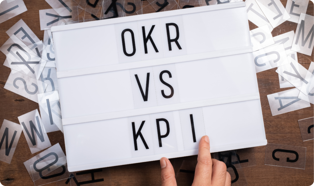 Uma placa luminosa branca de cinema exibe as palavras "OKR", "VS" e "KPI" em três linhas diferentes, escritas em letras pretas simples. Uma mão humana toca a letra "I" na base da placa, que está sobre uma mesa de madeira cercada por letras soltas espalhadas.