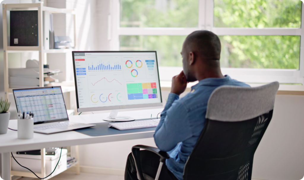 Visão traseira de um homem sentado em uma cadeira de escritório ergonômica, olhando para um grande monitor de computador que exibe um dashboard de Business Intelligence com diversos gráficos coloridos, mapas de calor e indicadores de performance. Um notebook aberto ao lado mostra uma planilha de dados detalhada.