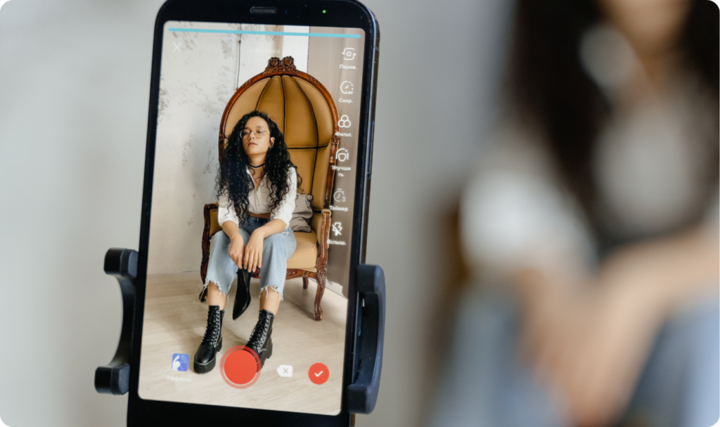 "Visão de um smartphone em um tripé capturando a gravação de um vídeo para o TikTok. Na tela, uma jovem modelo com óculos e cabelos cacheados posa em uma poltrona clássica, vestindo jeans e botas pretas. A interface do aplicativo exibe ícones de filtros e o botão de gravação vermelho, simbolizando o futuro das marcas no social commerce e a profissionalização dos criadores de conteúdo