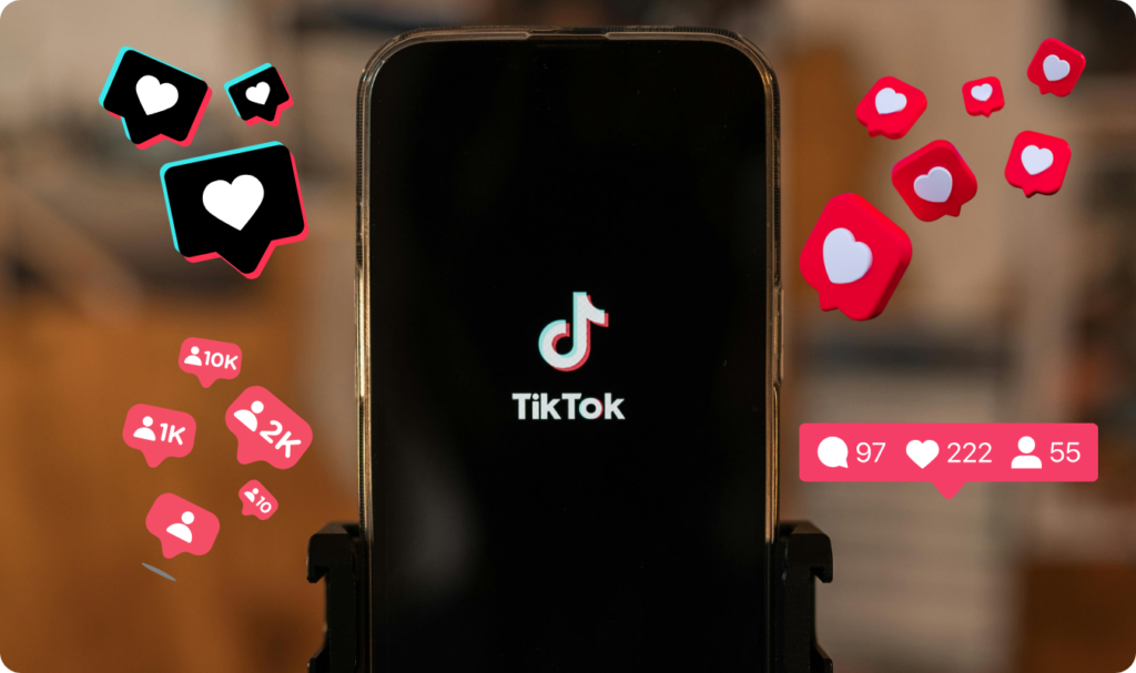 Fotografia em close-up de um smartphone exibindo o logotipo do TikTok em uma tela preta. Ao redor do aparelho, flutuam ícones de notificações estilizados em 3D, incluindo corações vermelhos de curtidas, balões de comentários e contadores de seguidores como '10K' e '2K'.