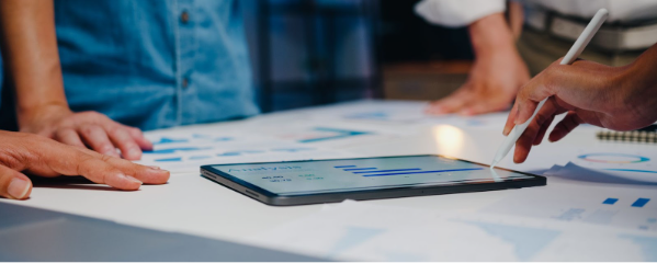 Equipe de profissionais em uma reunião de planejamento estratégico ao redor de uma mesa branca. No centro, um tablet exibe gráficos de análise de desempenho, enquanto uma mão utiliza uma caneta digital para destacar métricas. Braços de outros participantes e papéis com relatórios complementam a cena de trabalho colaborativo e tomada de decisão