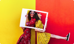 Duas mulheres sorridentes segurando uma moldura branca em frente a um fundo vibrante dividido entre amarelo e vermelho, representando a entrega de conteúdo e o alcance do algoritmo do Instagram.