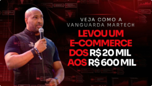 Um homem negro com barba curta, vestindo um colete preto com o logotipo da Vanguarda Martech, segura um microfone e fala durante uma apresentação. Ao lado dele, um texto em destaque sobre um fundo tecnológico vermelho escuro diz: "Veja como a Vanguarda Martech levou um e-commerce dos R$ 20 mil aos R$ 600 mil". O fundo da imagem contém gráficos e fluxogramas digitais de funis de vendas e métricas de marketing.