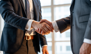 Close-up de dois executivos em ternos cinza escuro e camisas brancas apertando as mãos em sinal de acordo e parceria. A iluminação é quente e natural, vinda de uma grande janela ao fundo, destacando os detalhes dos punhos com abotoaduras e a firmeza do gesto. A imagem representa o sucesso em vendas, o fechamento de contratos e a consolidação de alianças estratégicas na Vanguarda Martech.
