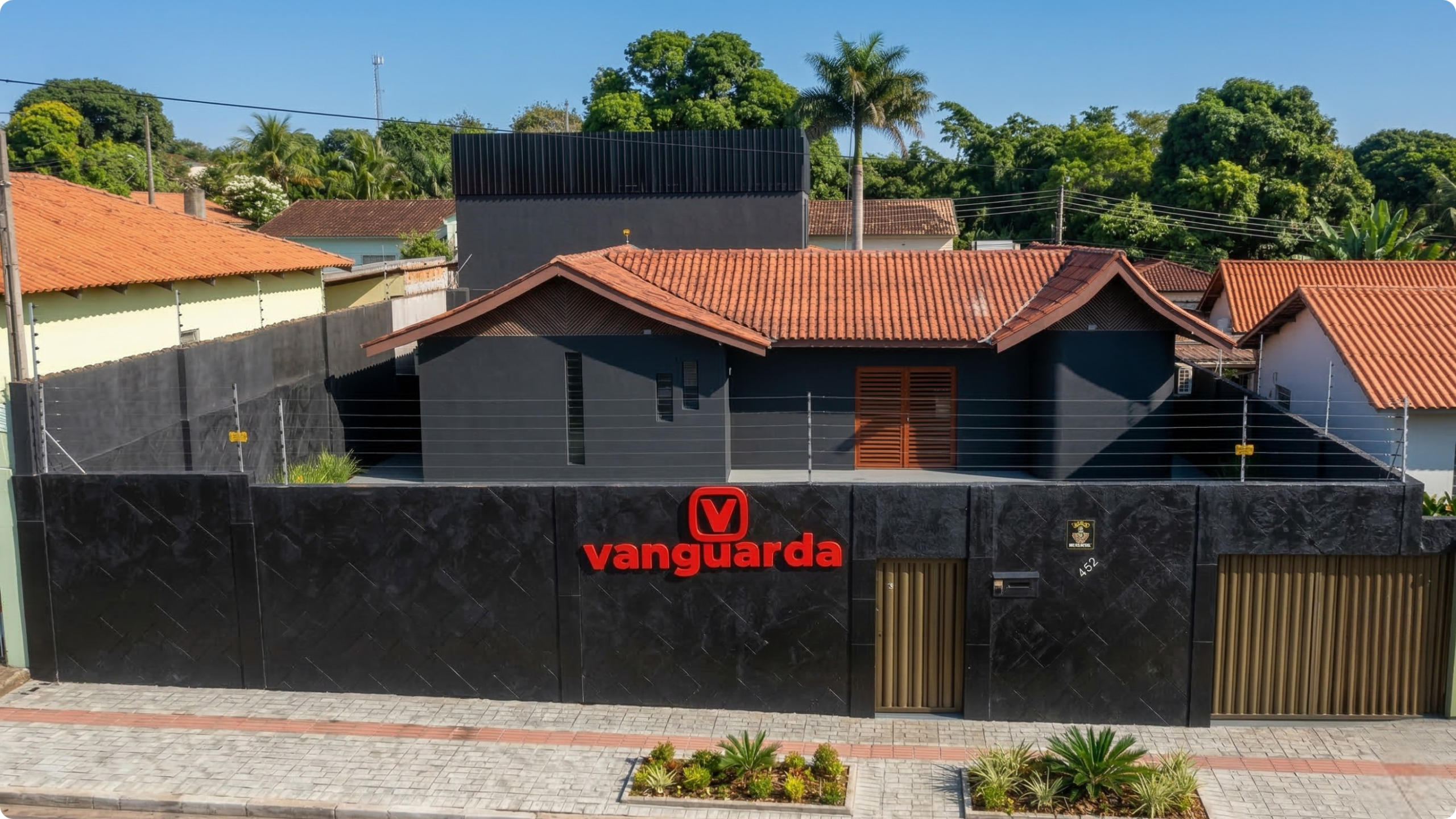 Uma fotografia aérea de alta resolução e ângulo frontal da fachada da Vanguarda Martech. O edifício possui paredes externas pintadas de preto fosco, com um logotipo vermelho destacado com a letra 'V' dentro de um quadrado e a palavra 'vanguarda' ao lado. A estrutura conta com um telhado de telhas cerâmicas alaranjadas e um anexo superior preto. À frente, há uma calçada de blocos intertravados cinza com pequenos canteiros de plantas ornamentais e um muro preto com detalhes em textura.