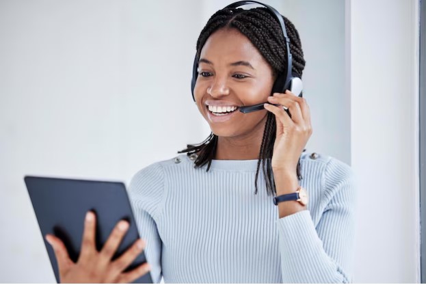 Mulher negra sorridente, com cabelos trançados, usando um headset preto e segurando um tablet. Ela veste uma blusa de tricô azul clara e olha para o dispositivo com uma expressão alegre e profissional.