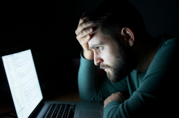 Homem barbudo sentado em um ambiente escuro, com expressão de cansaço ou intensa concentração, olhando para a tela de um notebook que ilumina seu rosto. Ele está com a mão na testa.